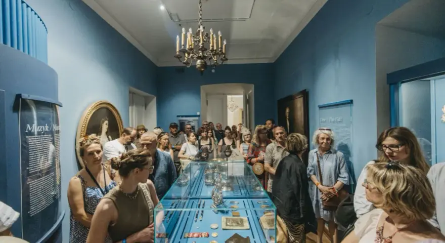 Grupa zwiedzających ogląda wystawę w muzeum. Wśród eksponatów w gablocie widać ozdobne przedmioty, a na ścianach obrazy i tablice informacyjne. Klasyczny wystrój sali z żyrandolem stanowi tło dla tego współczesnego zainteresowania historią.