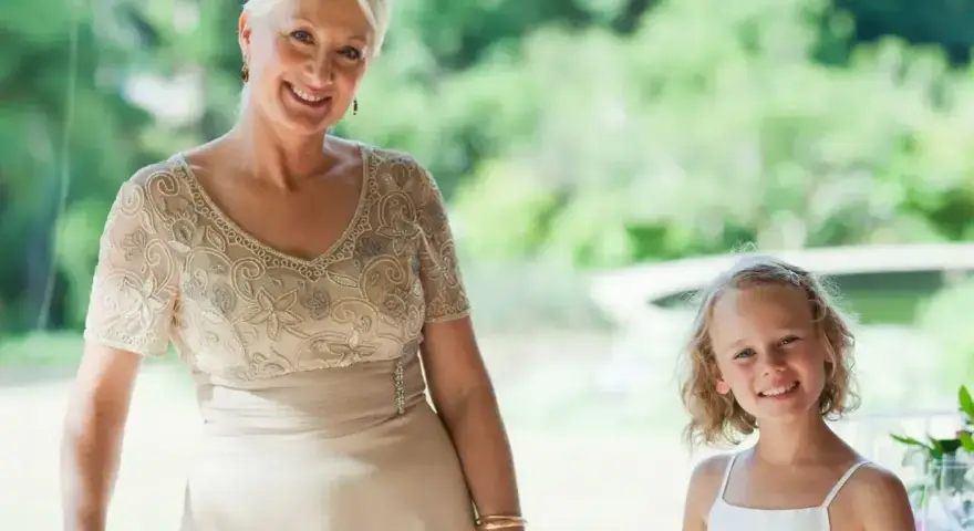 Elegancka stylizacja na komunię dla babci. Uśmiechnięta kobieta w beżowej sukni z haftem i dziewczynka w bieli.
