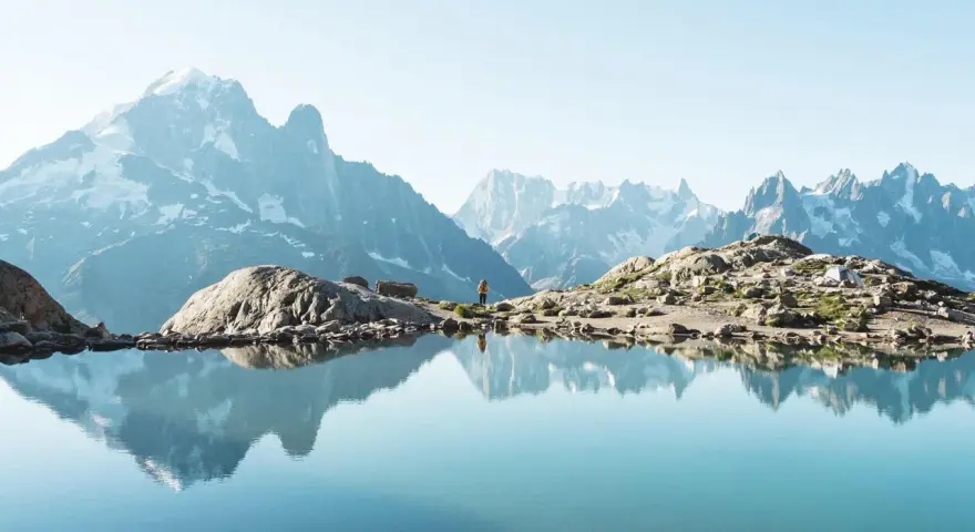 Un lac alpin reflète des montagnes majestueuses, un aperçu des plus belles randonnées de France. Une personne se tient sur la rive rocheuse.