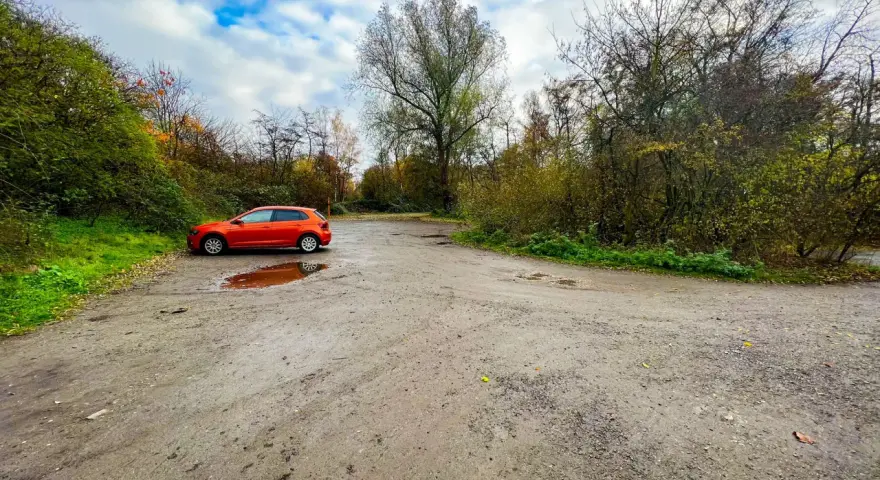 Ein orangefarbener VW Polo parkt auf einem matschigen Parkplatz am Rande eines Waldes. Die Halde Haniel Parken ist von Bäumen und Herbstlaub umgeben.