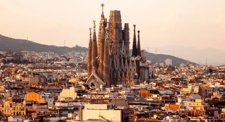 Vue panoramique de Barcelone au coucher du soleil, avec la Sagrada Familia au centre. Les travaux de la Sagrada Familia continuent, sa silhouette imposante se détachant sur le ciel orangé.