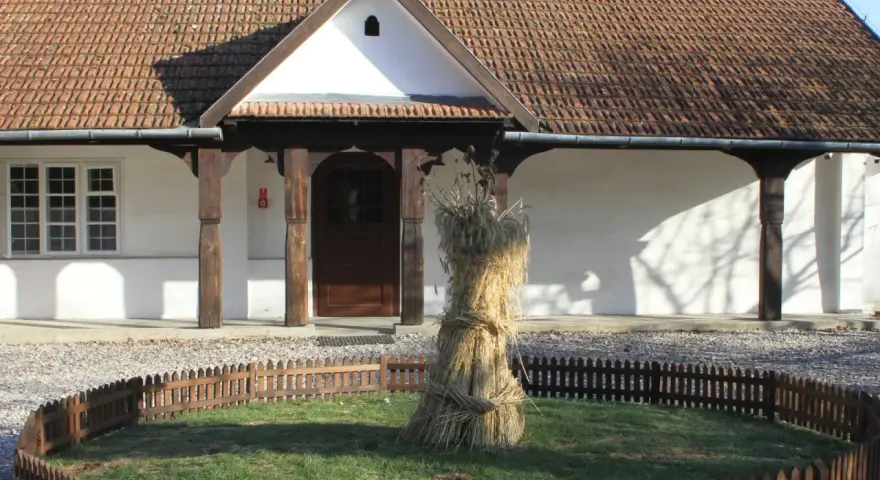 Bronowicka chata symbol: snop zboża przed tradycyjnym domem z drewnianymi elementami i ceramicznym dachem.