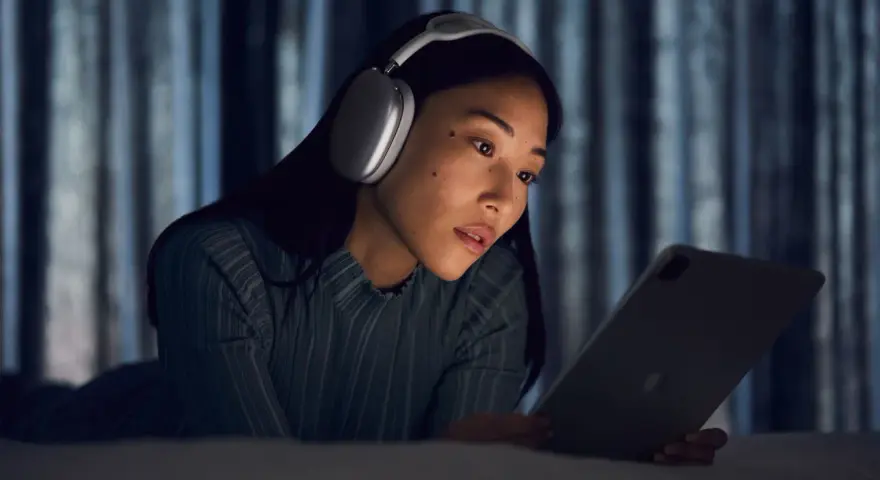 Kobieta w słuchawkach AirPods Max leży na łóżku i patrzy na tablet.
