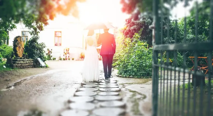 Para młoda idzie pod parasolem przez kamienną ścieżkę. W tle biały budynek i zielony ogród. To piękna brama weselna.