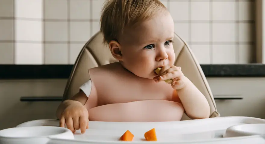 Małe dziecko w różowym śliniaku próbuje kawałek marchewki. Na tacce leżą kolejne pomarańczowe kawałki.