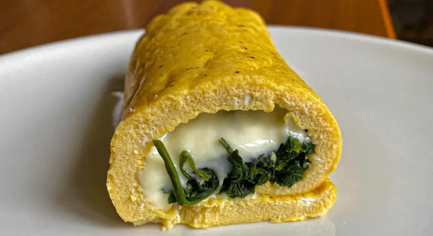 Rotolo di frittata dorato, ripieno di spinaci e formaggio filante, servito su un piatto bianco.