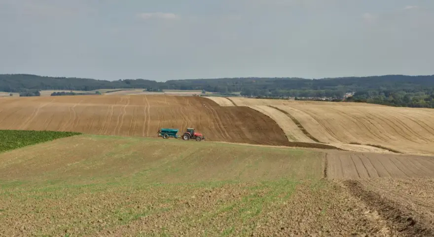 Czerwony traktor z przyczepą nawozi pole, przygotowując je do dalszej rekultywacji. W tle widać kolejne pola i las.