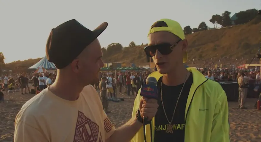 Dwóch mężczyzn na plaży, jeden z mikrofonem Red Bull, w tle tłum ludzi i scena.