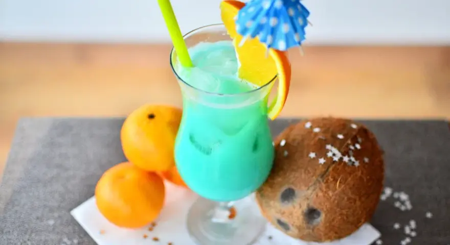 Orzeźwiający drink z sokiem ananasowym, ozdobiony plasterkiem pomarańczy i parasolką. Obok leżą pomarańcze i kokos.