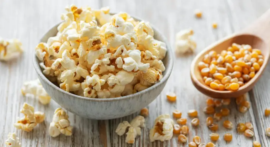 Schüssel mit frisch zubereitetem Popcorn im Topf, daneben eine Holzschaufel voller Maiskörner.