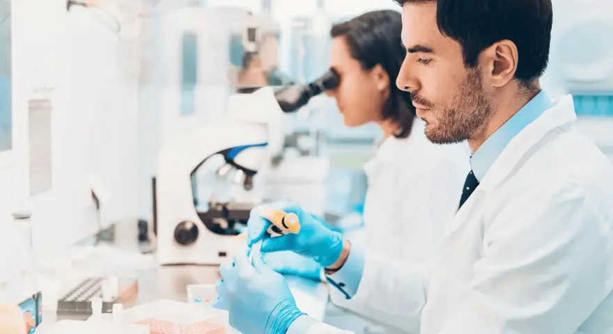 Ricerca di offerte di lavoro tecnico di laboratorio biomedico: due scienziati lavorano con provette e microscopio in un laboratorio moderno.