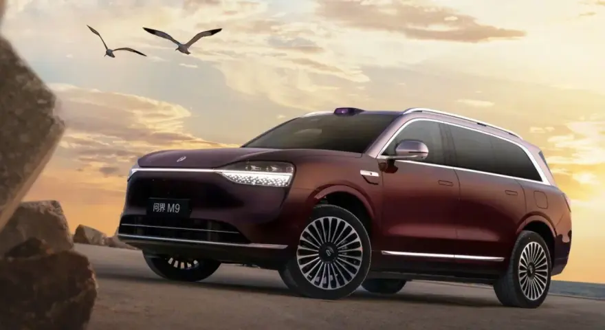 Ciemnoczerwony SUV Aito M9 na tle zachodzącego słońca, z ptakami w locie.