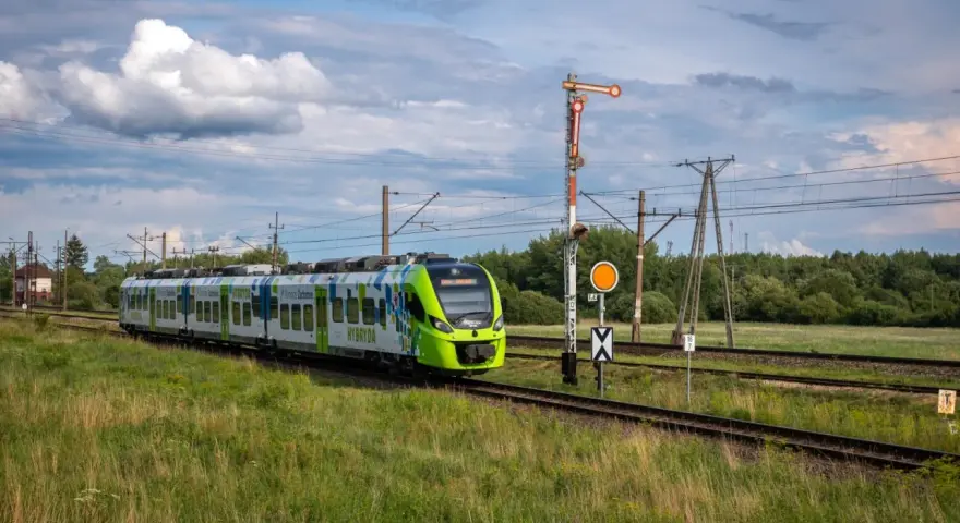 Zielony pociąg hybrydowy PKP jedzie po torach wśród zielonych pól. W tle las i chmury.