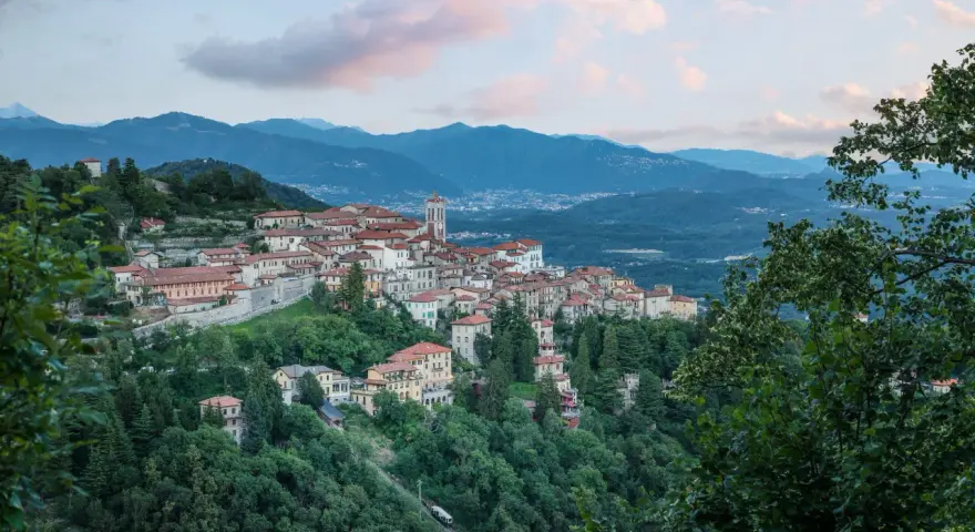 Borgo pittoresco su un colle, con case dai tetti rossi e un campanile, incastonato tra montagne e alberi. Via Francisca del Lucomagno, un incanto.