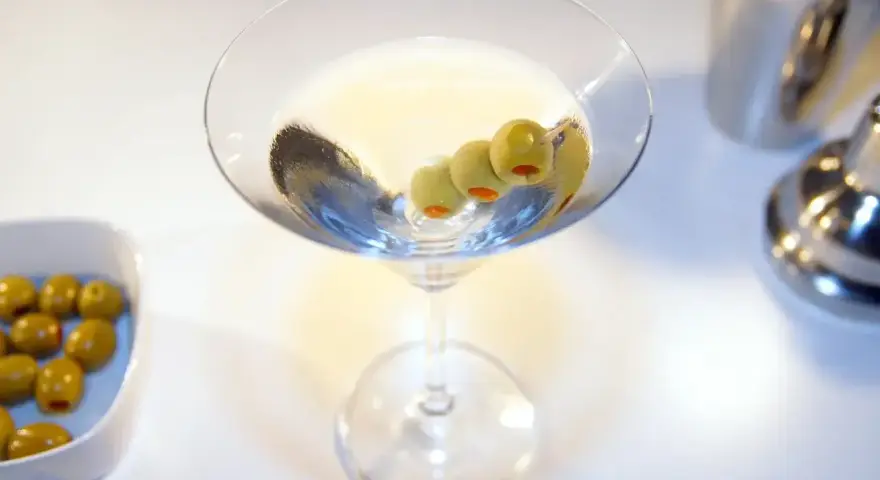 Elegancki drink z martini, ozdobiony trzema oliwkami, czeka na spróbowanie. Obok miseczka z oliwkami i shaker.