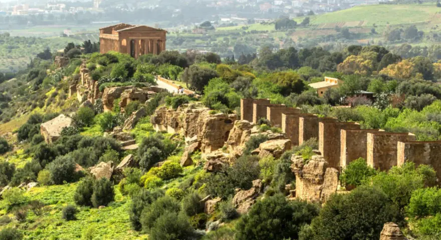 Ancient Greek temples stand proudly on a ridge in the Valley of the Temples, Sicily, surrounded by lush greenery and a distant town.