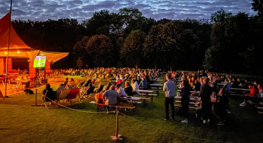 Menschen genießen ein Fußballspiel beim Public Viewing in Berlin unter einem dramatischen Abendhimmel.