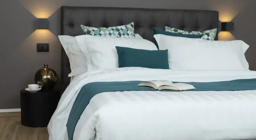Letto rifatto con lenzuola bianche e copriletto turchese. Un libro aperto è posato al centro, pronto per essere letto. Ecco come si mette il lenzuolo di sopra.
