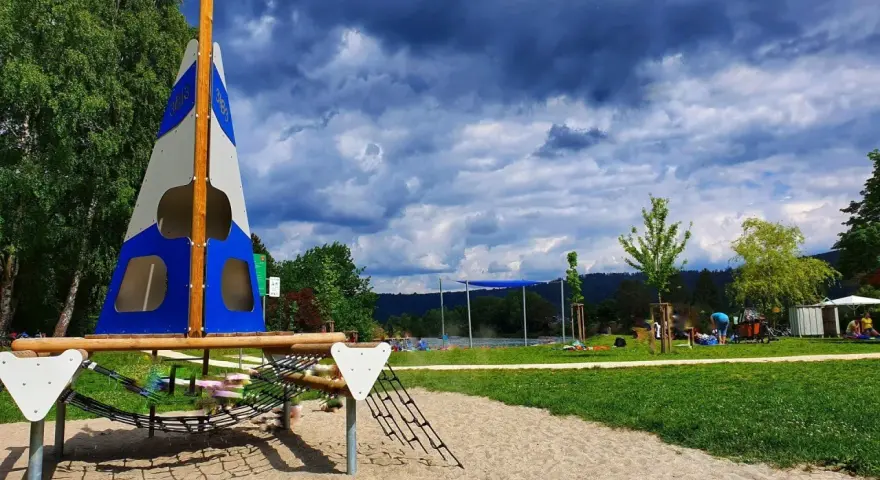 Spielplatz am Badesee Ludwigsburg mit Segel-Klettergerüst und Sandkasten. Im Hintergrund der See und Menschen.