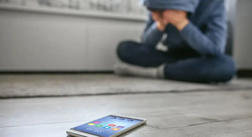Osoba w kapturze siedzi na podłodze, ukrywając twarz w dłoniach. Obok leży telefon, symbolizujący izolację i dystymię.