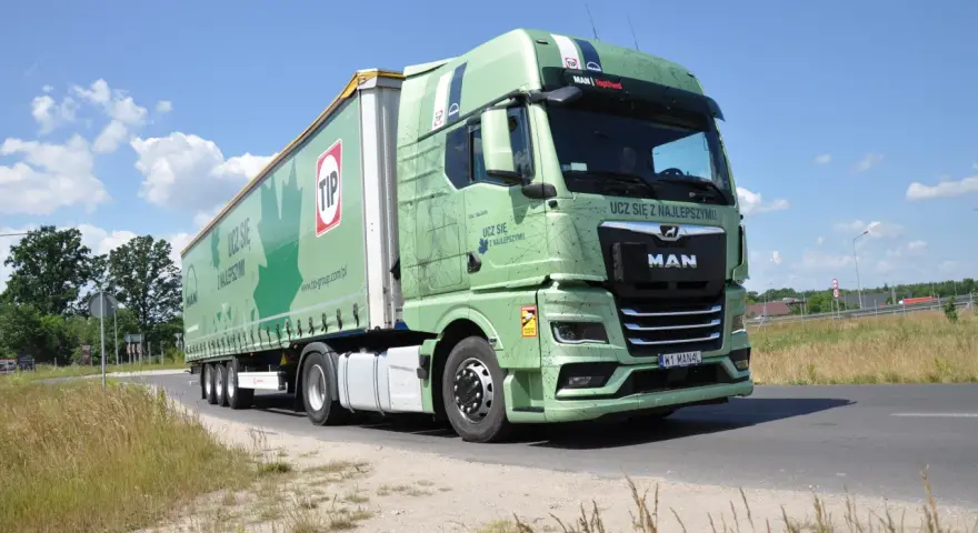 Zielona ciężarówka MAN TGX z naczepą TIP jedzie drogą. Na naczepie widnieje napis "Ucz się z najlepszymi!".