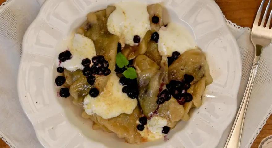 Knedle ze śliwkami czy pierogi z jagodami? Na talerzu pyszne pierogi z jagodami, polane śmietaną i posypane świeżymi owocami.