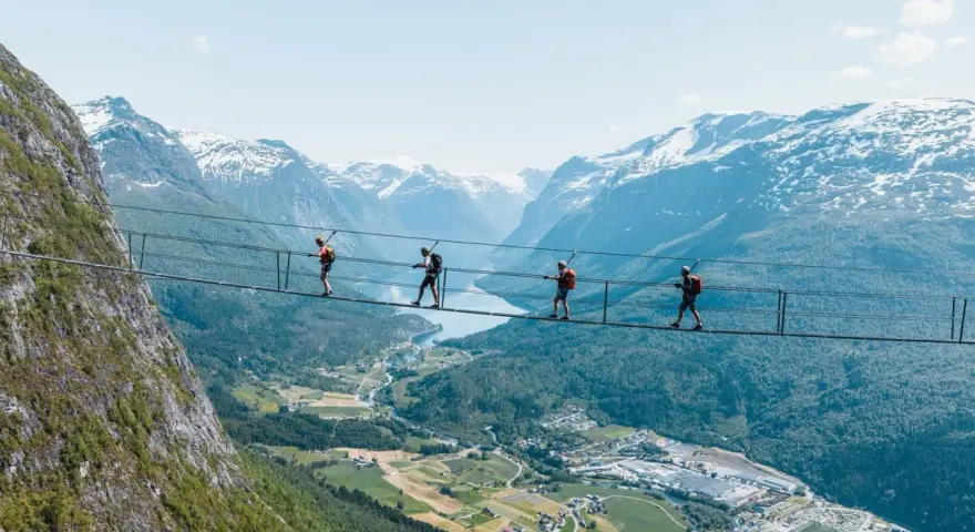 Fyra personer går på en hängbro vid Loen via ferrata, med en fantastisk utsikt över fjord och snöklädda berg.