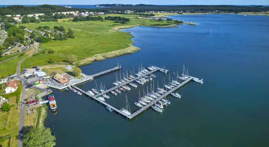 Der Hafen Sellin ist voller Segelboote, die auf dem ruhigen Wasser liegen. Im Hintergrund erstreckt sich eine grüne Landschaft mit Häusern und Bäumen.