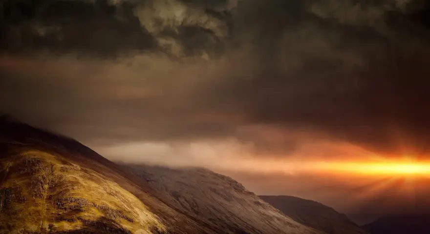 Dramatic sunburst pierces dark clouds over a rugged mountain range, a powerful display of the sovereignty of God.