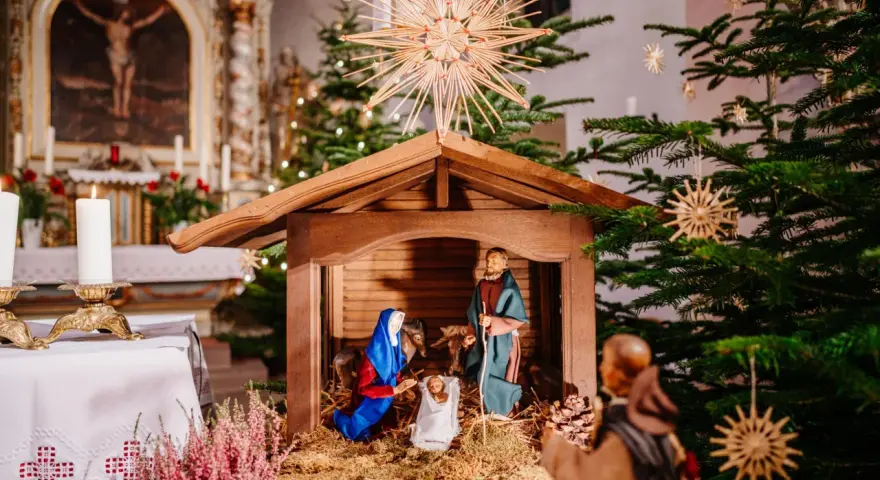 Weihnachtskrippe in einer Kirche, die die Geburt Jesu darstellt. Maria und Josef knien vor dem Kind, während im Hintergrund ein Weihnachtsbaum und Kerzen leuchten. So feiert man Weihnachten.