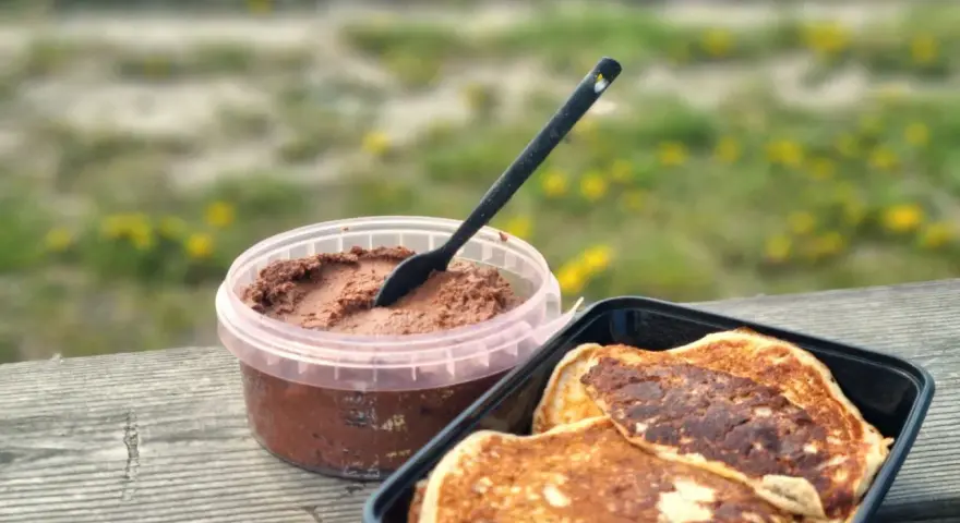 Färdiga proteinpannkakor i en svart behållare bredvid en burk med chokladpålägg, redo att avnjutas utomhus.