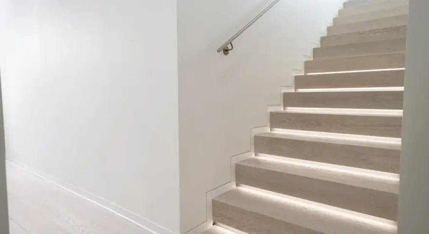 Scala moderna con gradini in legno illuminati e un discreto profilo battiscopa filo muro che ne esalta la pulizia delle linee.