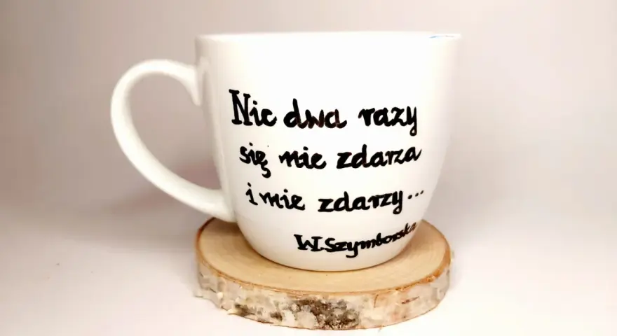 Biały kubek z cytatem Wisławy Szymborskiej: "Nic dwa razy się nie zdarza i nie zdarzy...". Wczoraj kiedy twoje imię ktoś wymówił przy mnie głośno wiersz.