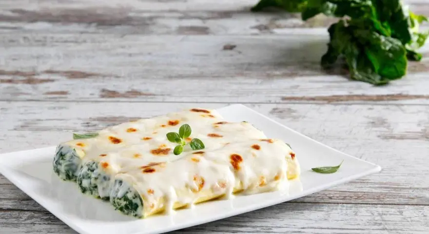 Cannelloni ricotta e spinaci fumanti, ricoperti di besciamella e gratinati, guarniti con un rametto di origano.
