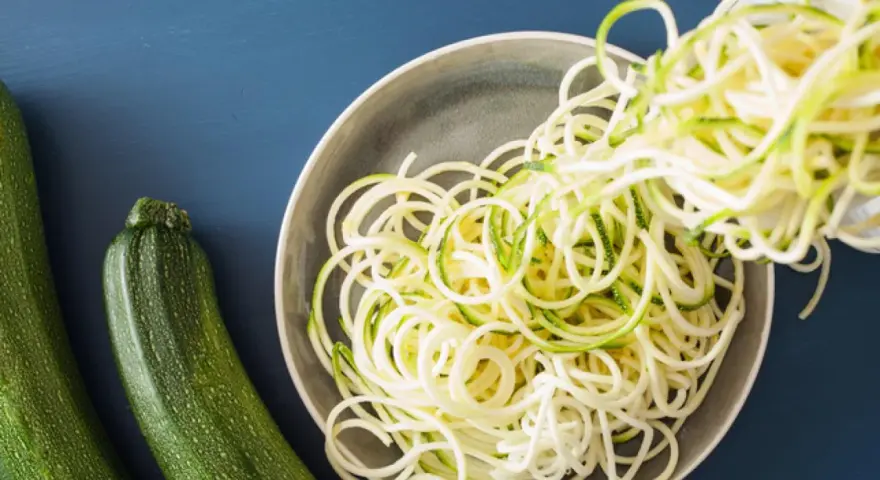 Espaguetis con calabacín recién hechos, listos para servir. Dos calabacines verdes reposan al lado de un plato lleno de fideos de calabacín.
