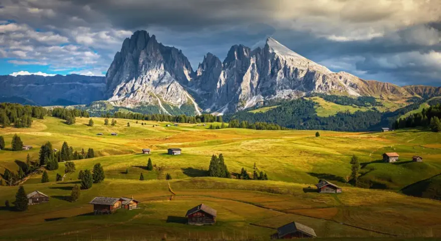 Un paesaggio alpino con un sasso lungo e imponente che domina un sasso piatto e verdeggiante, punteggiato da casette di legno.