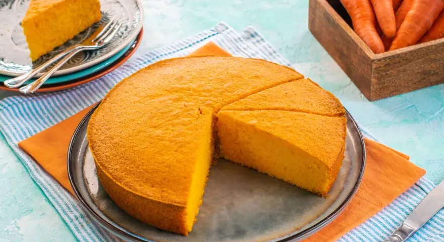 Una fetta di torta di carote sofficissima, appena tagliata da una torta intera su un piatto, con carote fresche in un cesto sullo sfondo.