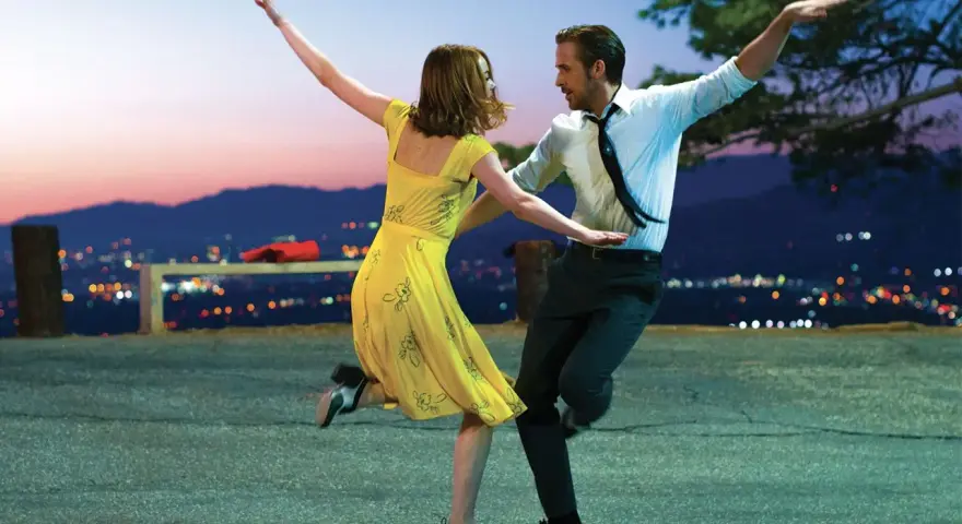Coppia balla al tramonto, immersa nella musica diegetica che accompagna la loro danza, con la città illuminata sullo sfondo.