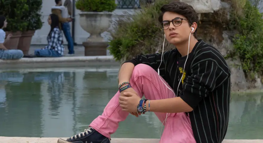 Il ragazzo dai pantaloni rosa, con occhiali e auricolari, ascolta musica seduto vicino a una fontana, mentre altre persone chiacchierano sullo sfondo.