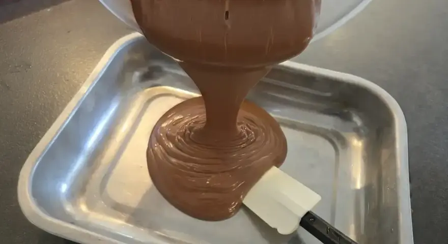 Une ganache au chocolat, inspirée par le Toblerone, est versée dans un plat métallique. Une spatule aide à l'étaler.