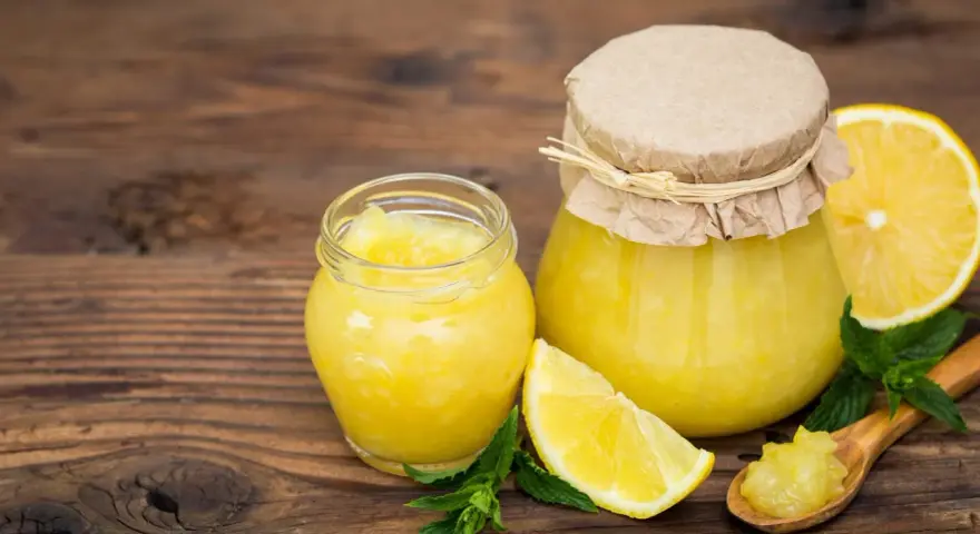 Due vasetti di marmellata di limoni, limoni freschi e foglie di menta su un tavolo di legno.