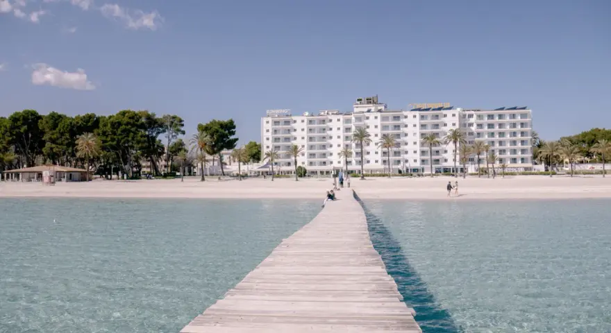 Drewniany pomost prowadzi do piaszczystej plaży Playa de Muro z białym hotelem i palmami w tle.