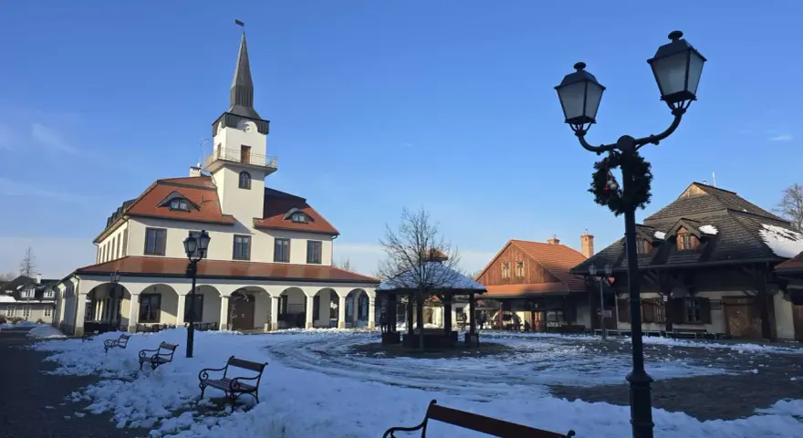 Zimowe miasteczko galicyjskie z ratuszem i karczmą, plac pokryty śniegiem, ławki i latarnie ozdobione świąteczną girlandą.