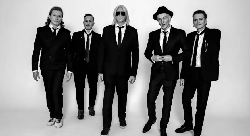 Fünf Männer in schwarzen Anzügen und Krawatten, die wie der titel von Def Leppard aussehen, posieren für ein Schwarz-Weiß-Foto.