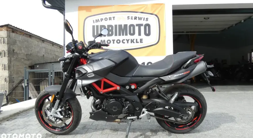 Czarny motocykl Aprilia Shiver 900 z czerwonym stelażem, zaparkowany przed salonem motocyklowym.