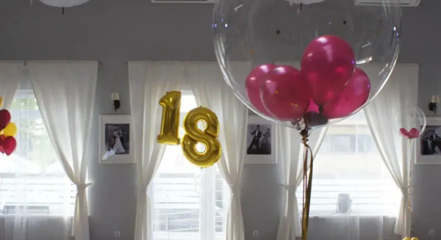 Złote balony z cyfrą 18 i przezroczyste balony z różowymi w środku ozdabiają salę. Balony na stołach tworzą radosną atmosferę.