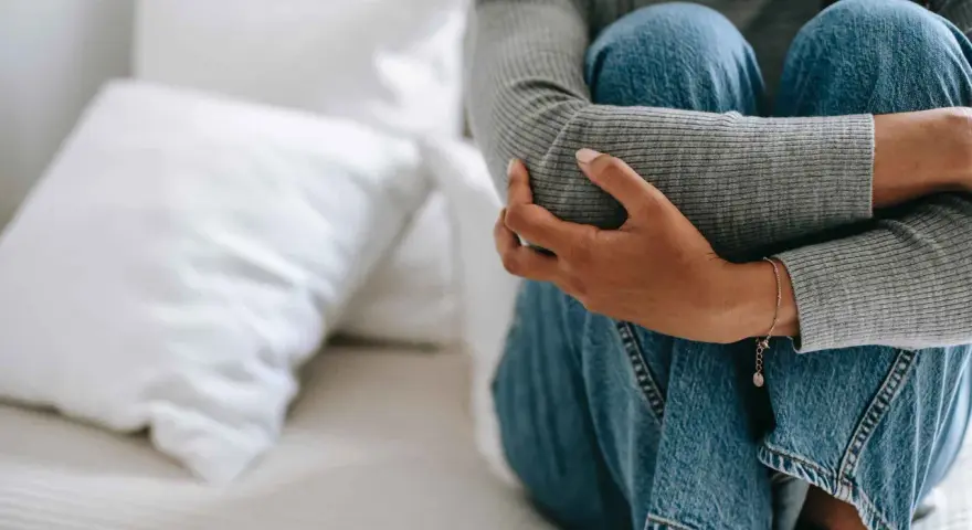 Person sitter hopkrupen på en säng, omfamnar sina knän. Klädd i jeans och grå tröja, utstrålar bilden extrem ångest.