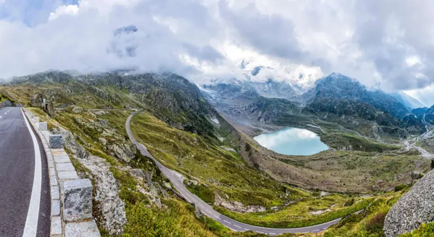 Strada tortuosa sul Passo San Lugano, con lago alpino azzurro e montagne nebbiose sullo sfondo.