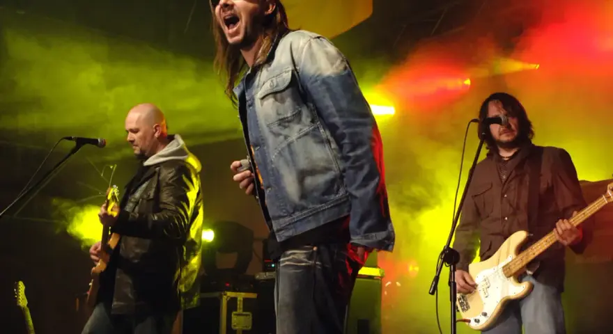 Energetyczne kapele punkowe z lat 80. na scenie, w dymie i kolorowych światłach. Wokalista krzyczy do mikrofonu, gitarzysta gra na basie.