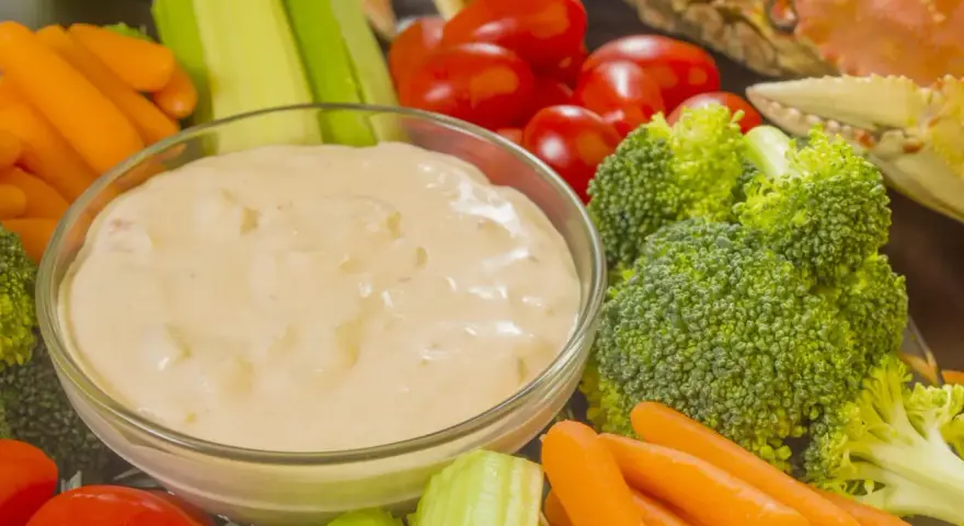 Un bol de sauce crudités crème fraîche entouré de carottes, céleri, tomates cerises et brocolis. Un crabe cuit est visible en arrière-plan.